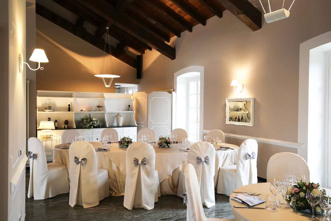 Sala ricevimenti con tavole apparecchiate, sedie con copridivani bianchi e travi in legno