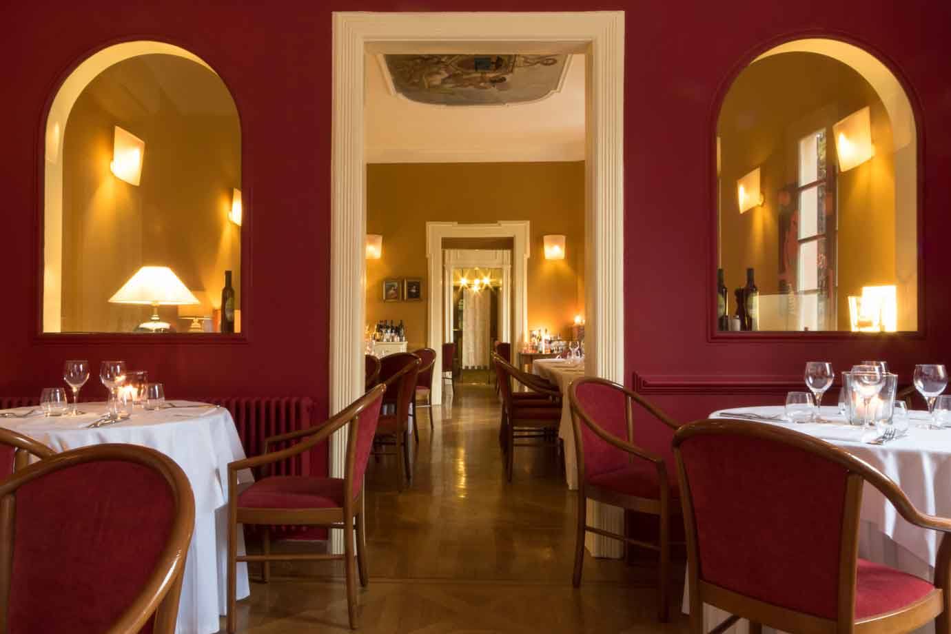 Sala da pranzo con pareti bordeaux, specchi ad arco, tavoli imbanditi e luce calda