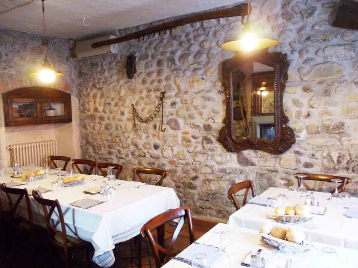 Sala di trattoria con tavoli apparecchiati, pareti in pietra, specchio antico e lampade accese