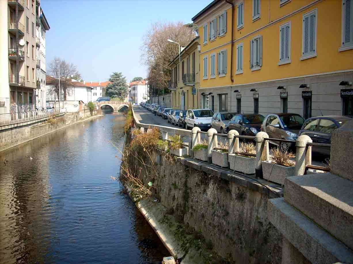 Auto parcheggiate accanto al canale Naviglio tra edifici gialli e un ponte
