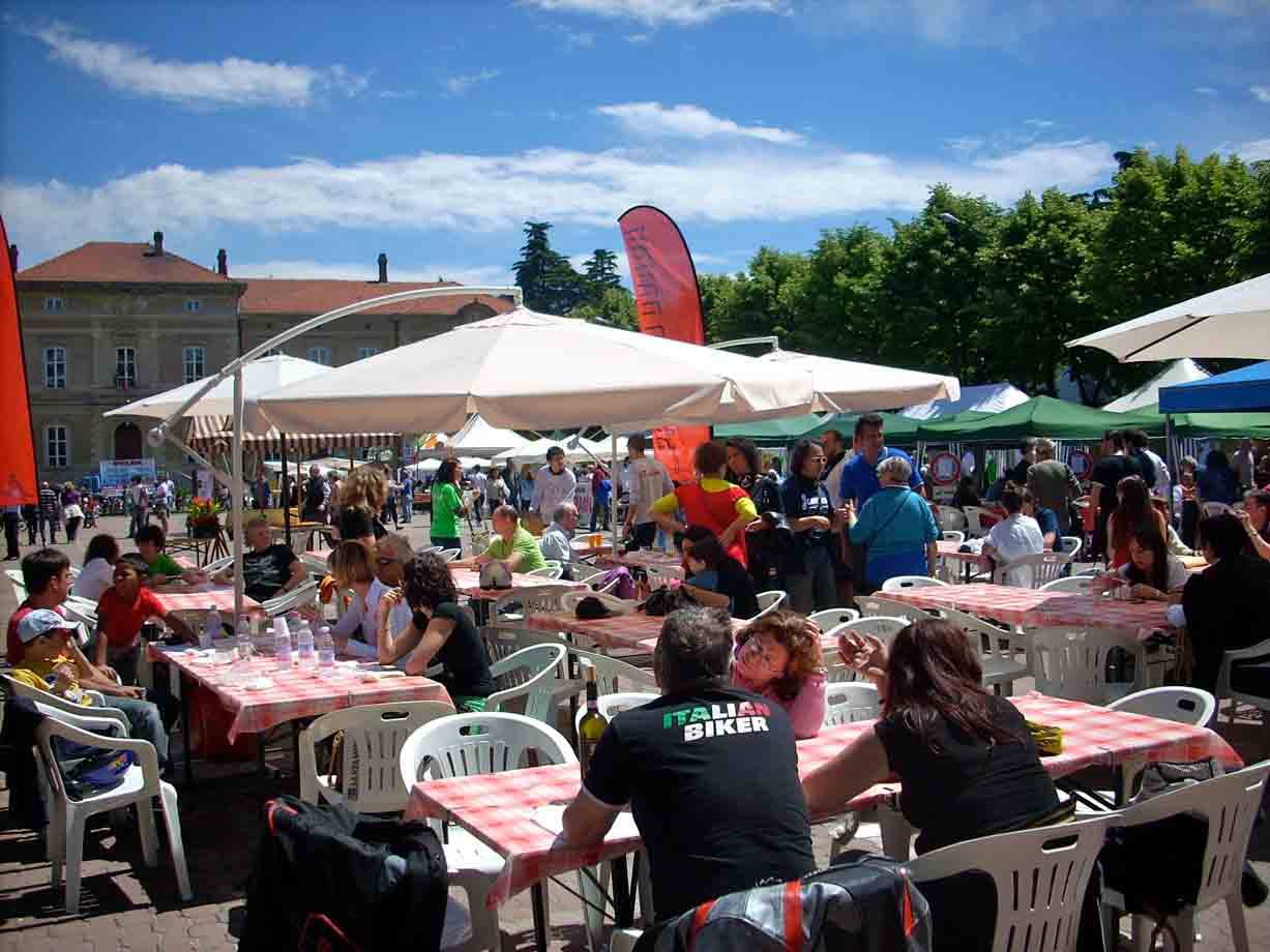 Tavoli con tovaglie a quadri sotto ombrelloni bianchi, folla in piazza soleggiata