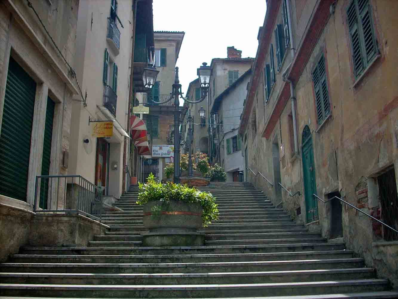 Scalinata in pietra tra case con persiane verdi, fiori e lampioni in centro