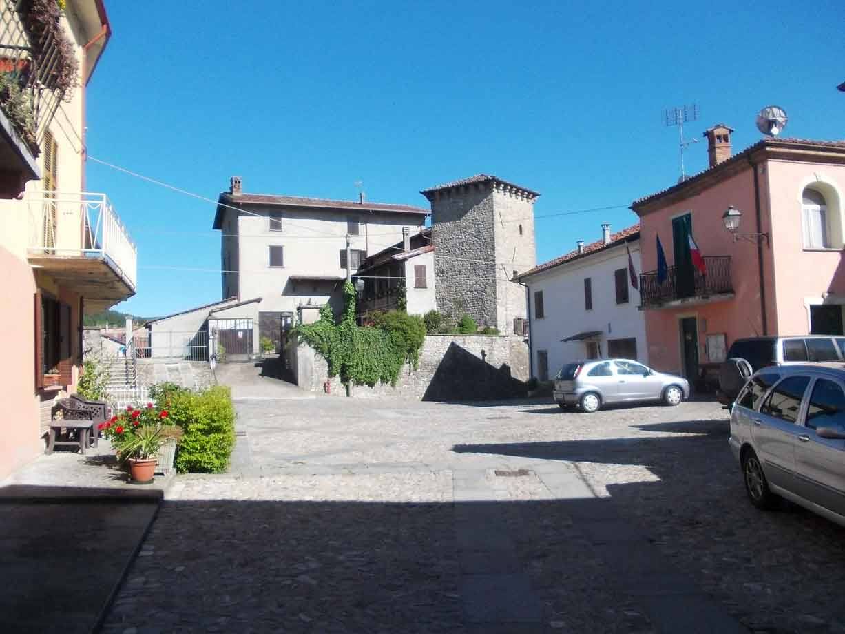 Piazzale in ciottoli con case colorate, torre di pietra e auto parcheggiate al sole