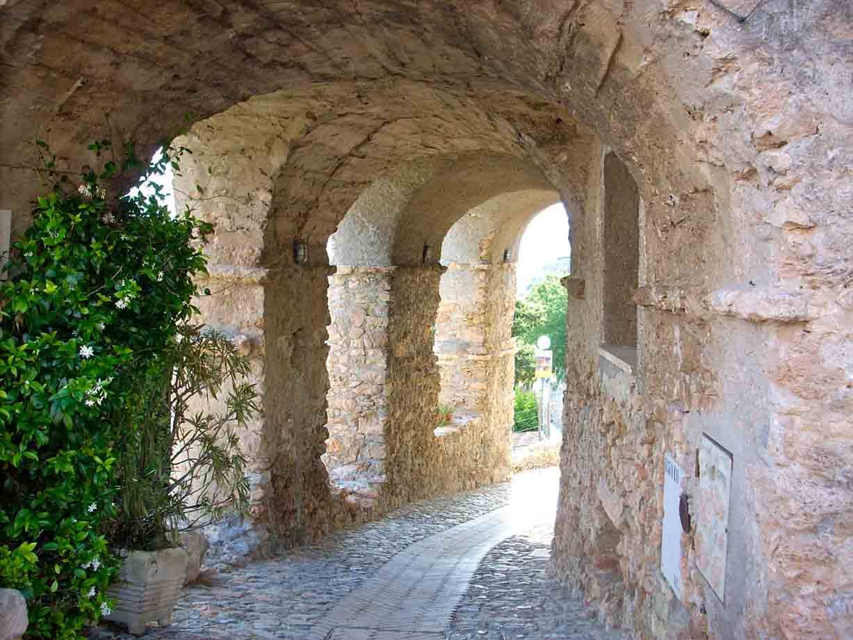 Portico di pietra con archi, piante e strada lastricata illuminata dal sole