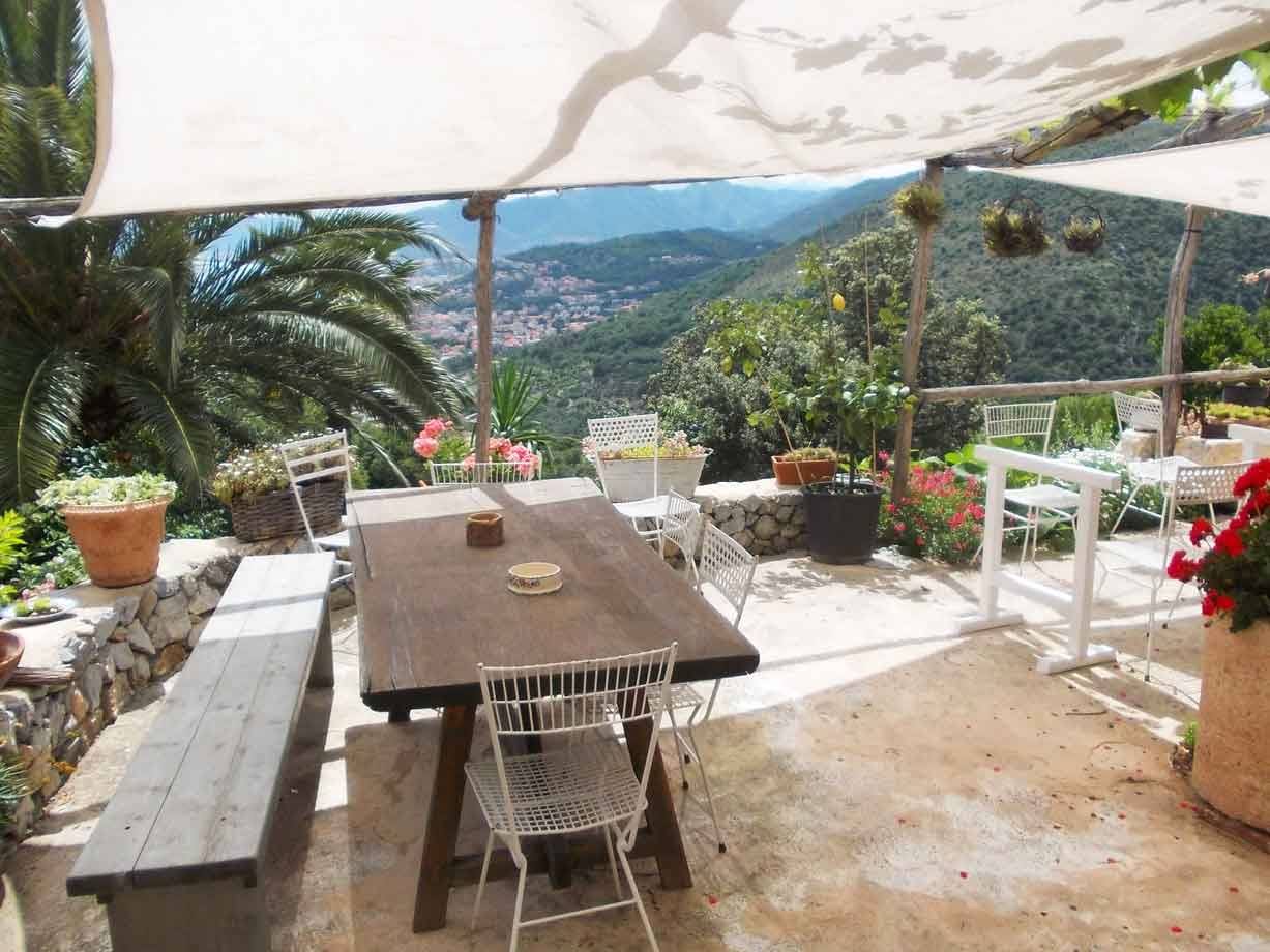 Tavolo in legno sotto teli ombreggianti, terrazza con vasi di fiori e montagne
