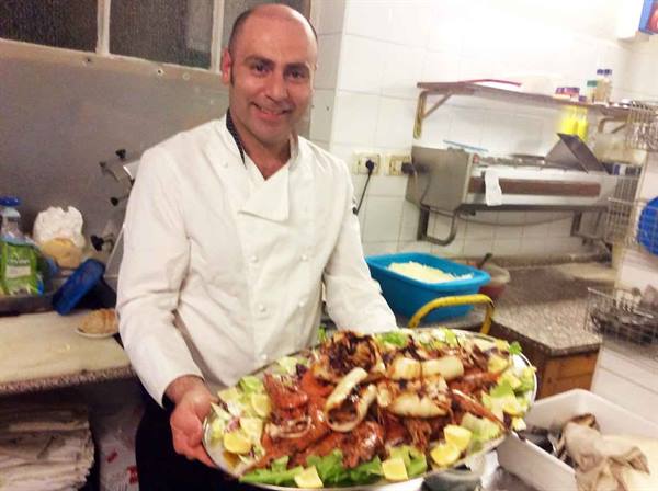 Smiling chef in kitchen presents a large plate of calamari and shrimp on lettuce