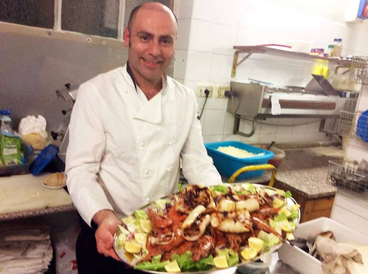Chef sorridente in cucina presenta un grande piatto di calamari e gamberi su insalata