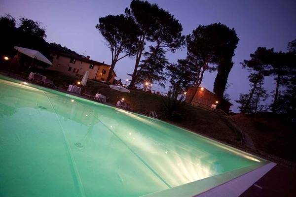 Illuminated pool at night beside a villa with umbrellas and pines