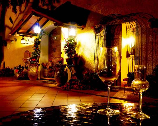 Wine glasses on table, stone courtyard lit by lamps, flowers and terracotta pots