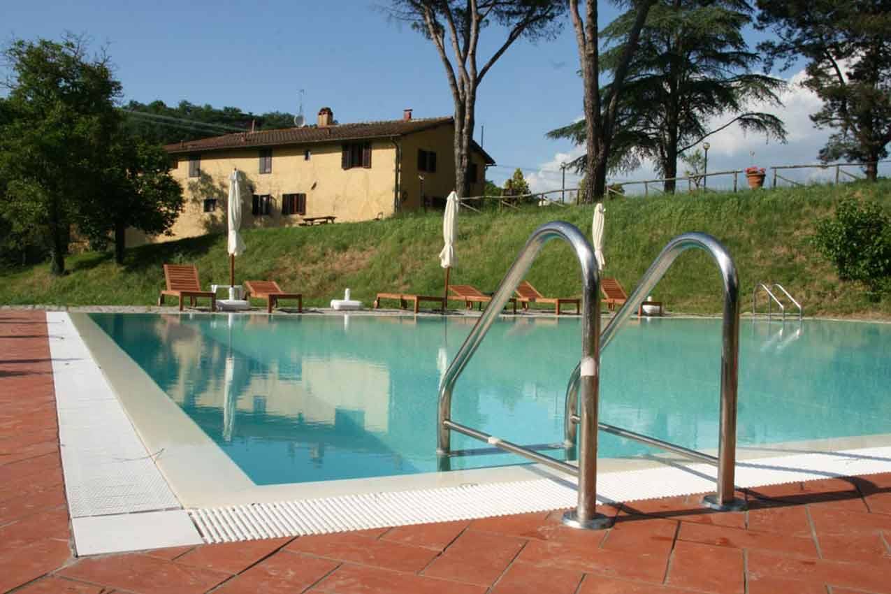 Piscina con scaletta in acciaio davanti a casale giallo tra alberi, ombrelloni e lettini