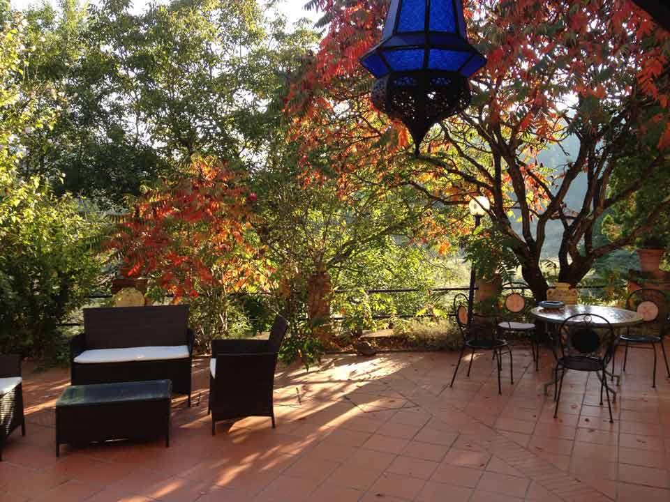 Terrazza in cotto con salotto di rattan, tavolo in ferro e lanterna blu tra alberi autunnali