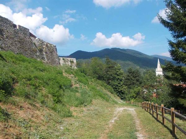 Stone walls along grassy path, white bell tower and mountains under cloudy sky