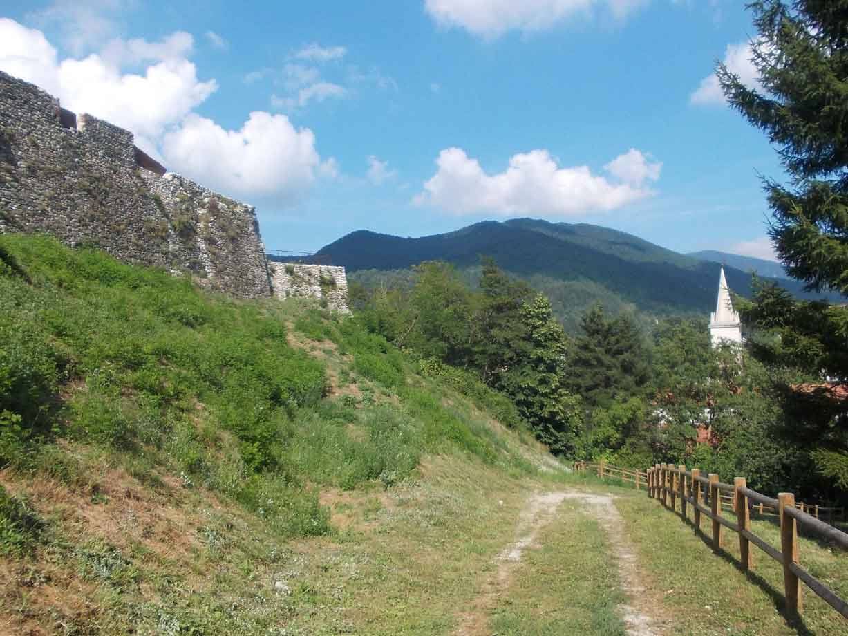 Mura in pietra lungo sentiero erboso, campanile bianco e monti sotto cielo nuvoloso