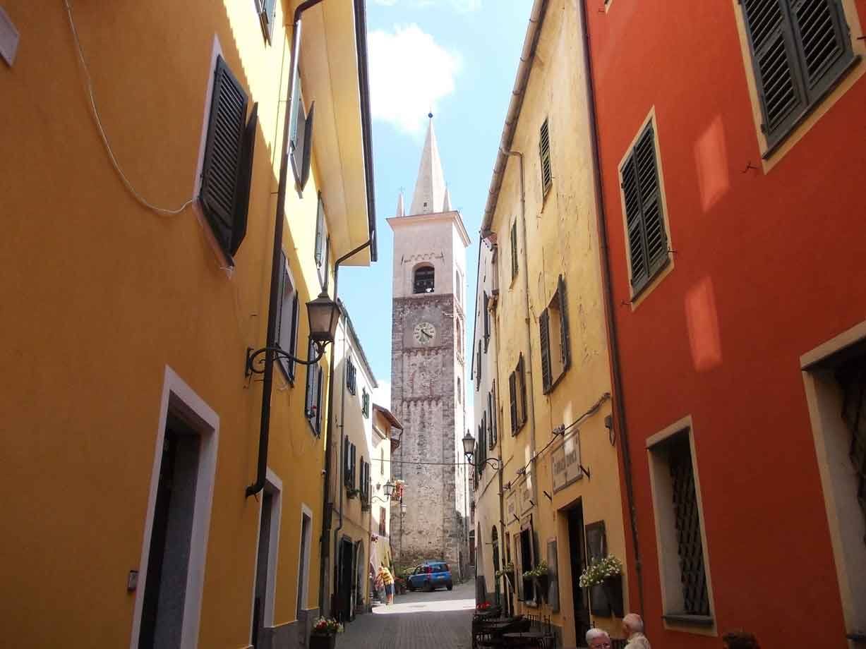 Vicolo tra case gialle e arancioni, campanile in pietra con orologio al fondo