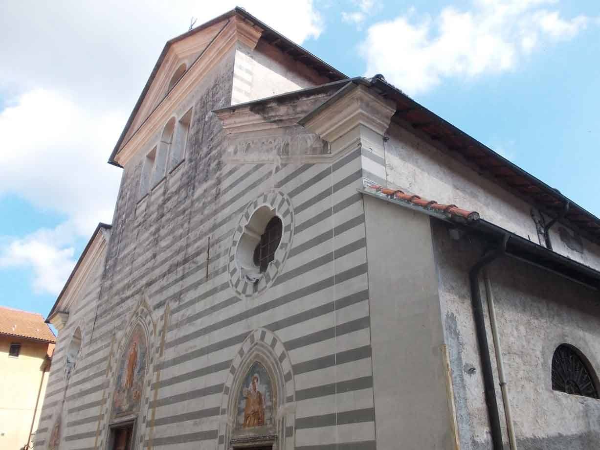 Facciata a bande grigie e bianche di chiesa medievale con occhio circolare e affreschi, cielo nuvoloso