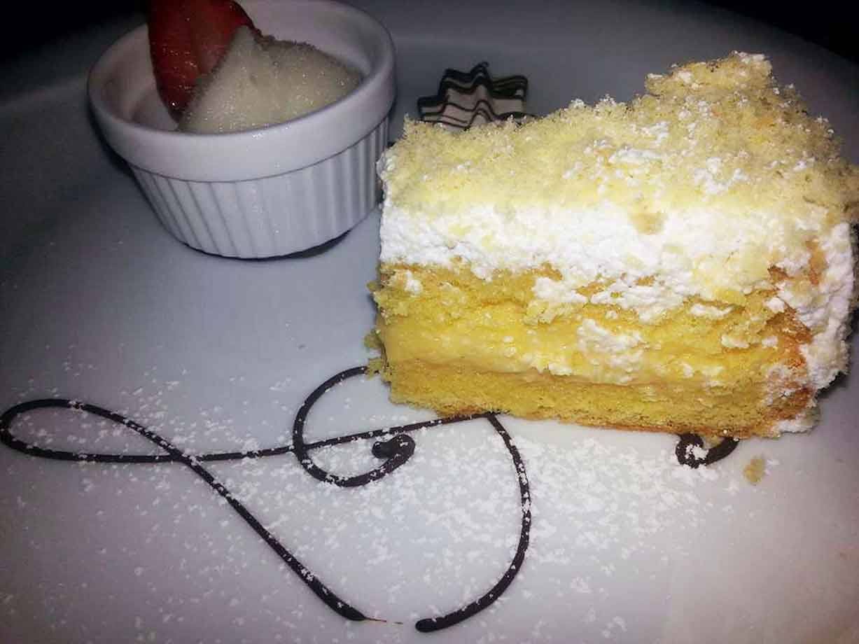 Fetta di torta mimosa con crema al piatto, accanto coppetta con fragola e panna