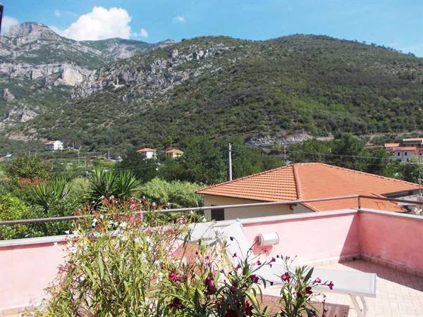 Pink terrace with flowers and sun lounger, terracotta rooftops beneath green mountains