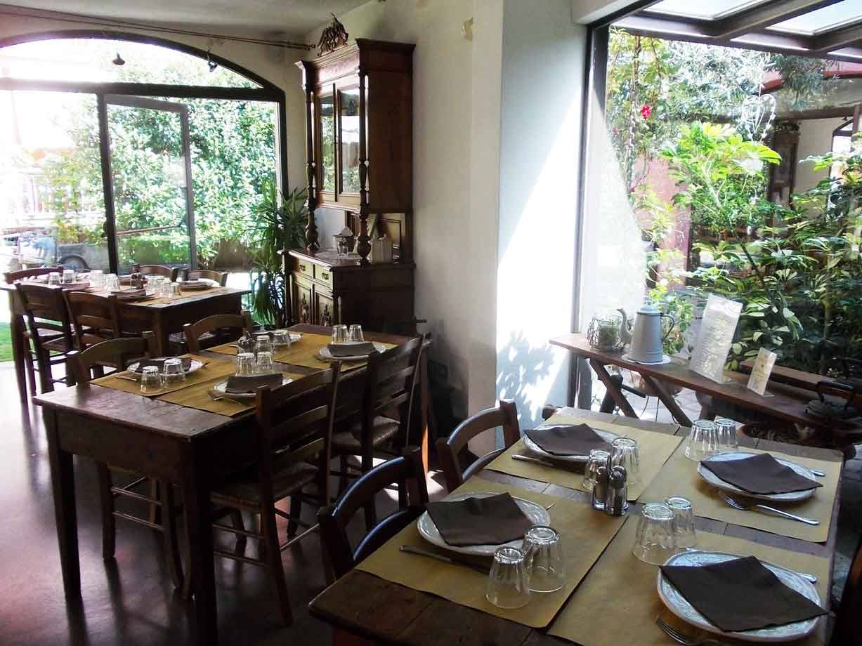 Sala di trattoria con tavoli in legno apparecchiati, credenza antica e vetrate sul giardino