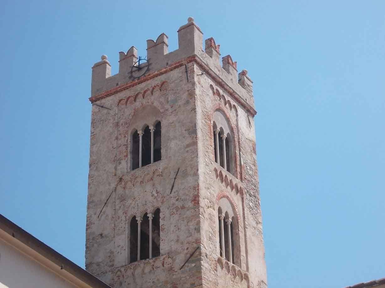 Campanile medievale in pietra e mattoni con bifore e merlatura, contro cielo azzurro