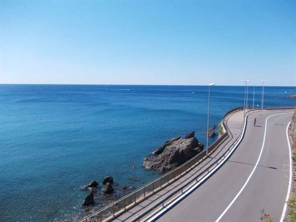 Coastal asphalt road curving above the rocks, lampposts and guardrail by the blue sea