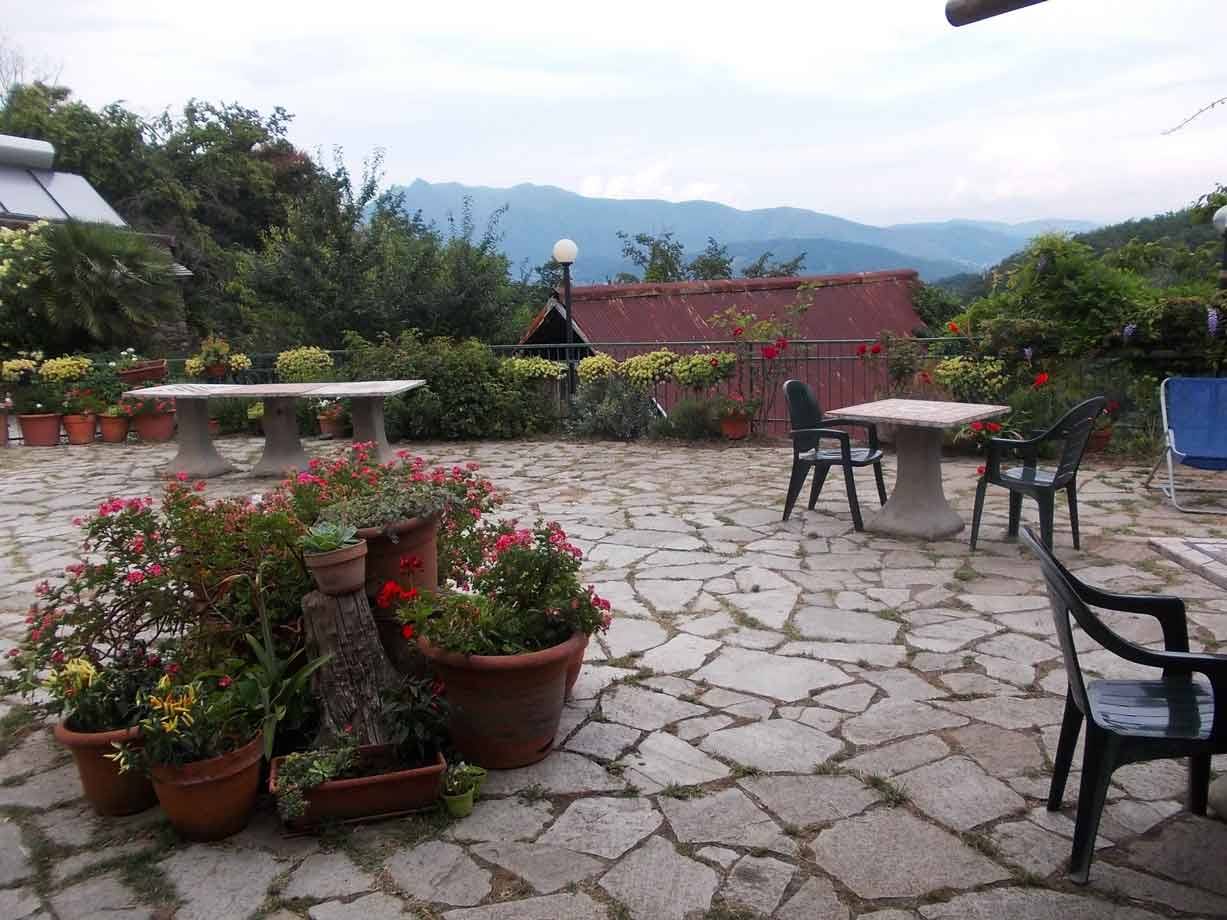 Terrazza in pietra con vasi di gerani, tavoli e sedie plastiche, monti nuvolosi