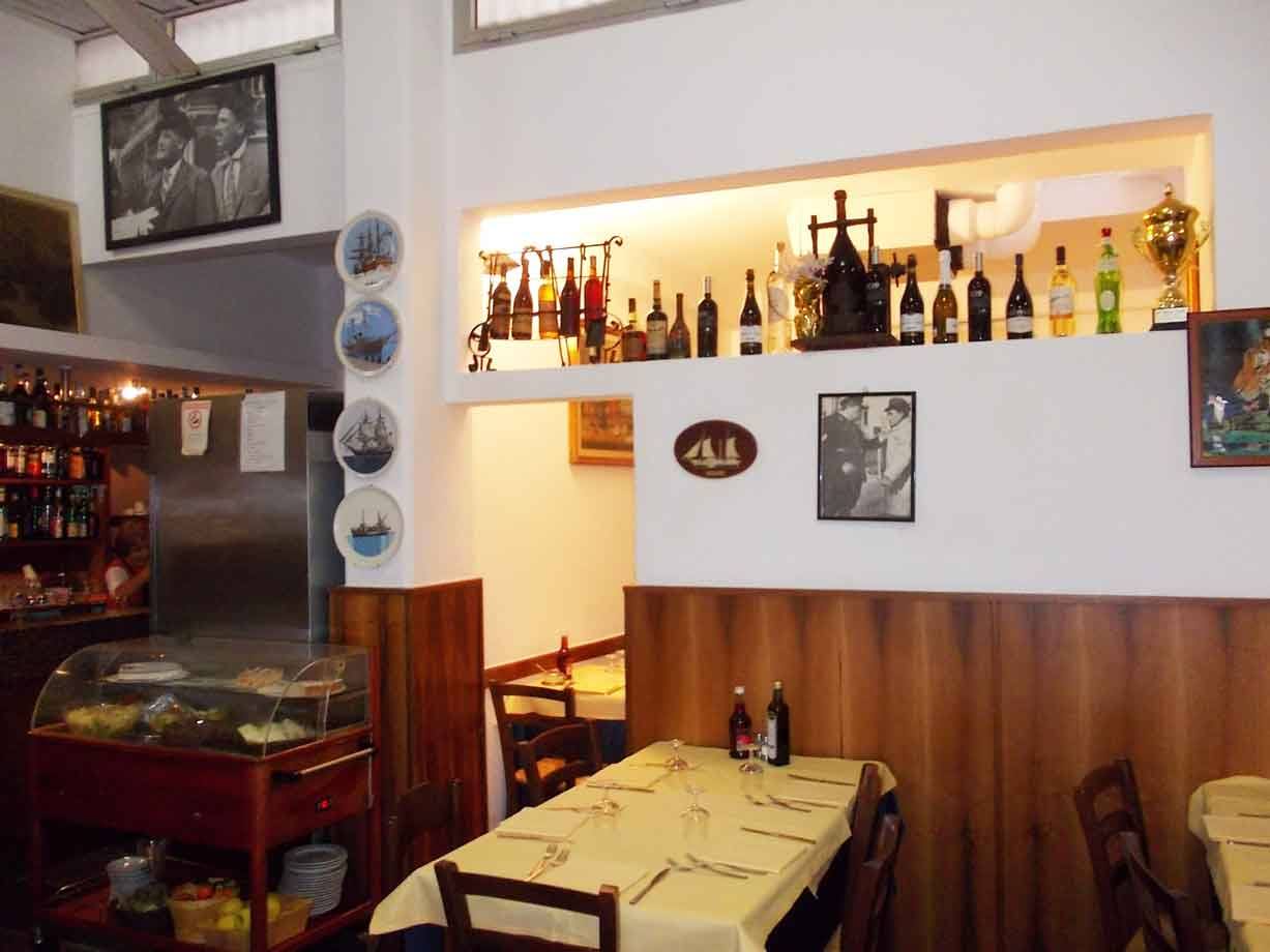 Sala di trattoria con tavoli apparecchiati, bottiglie illuminate e piatti decorati alle pareti