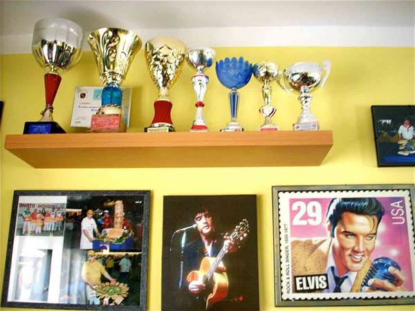 Silver trophies on wooden shelf above framed photos and an Elvis stamp
