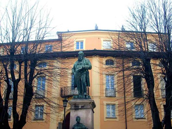 Bronze statue on pedestal before ochre building with windows and bare trees