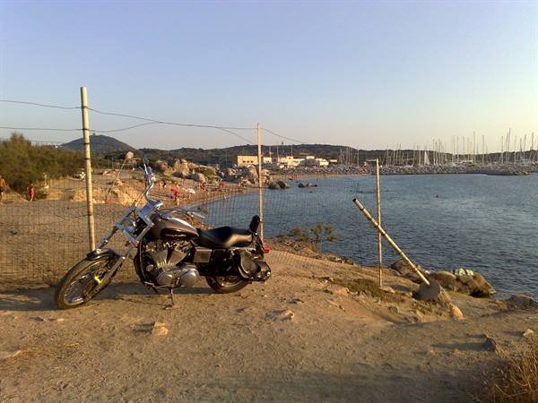 Harley motorcycle parked on sand near the fence, marina in the background