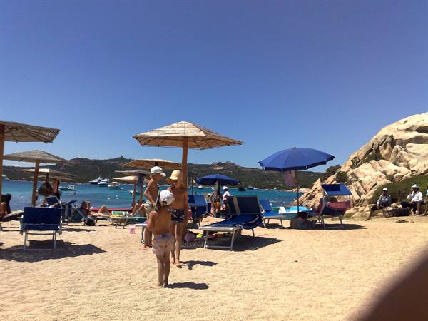 Children under straw parasols on sand, blue umbrellas and rocks, surely Spargi