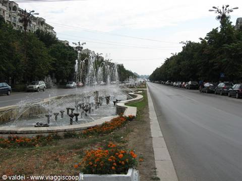 Stone fountains along a tree-lined boulevard in Bucharest with parked cars