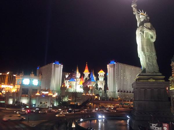 Replica Statue of Liberty in front of illuminated Excalibur castle, nighttime traffic in Las Vegas