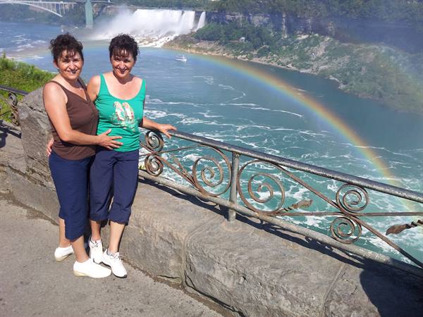 Two smiling women by an iron railing in front of Niagara Falls with a rainbow