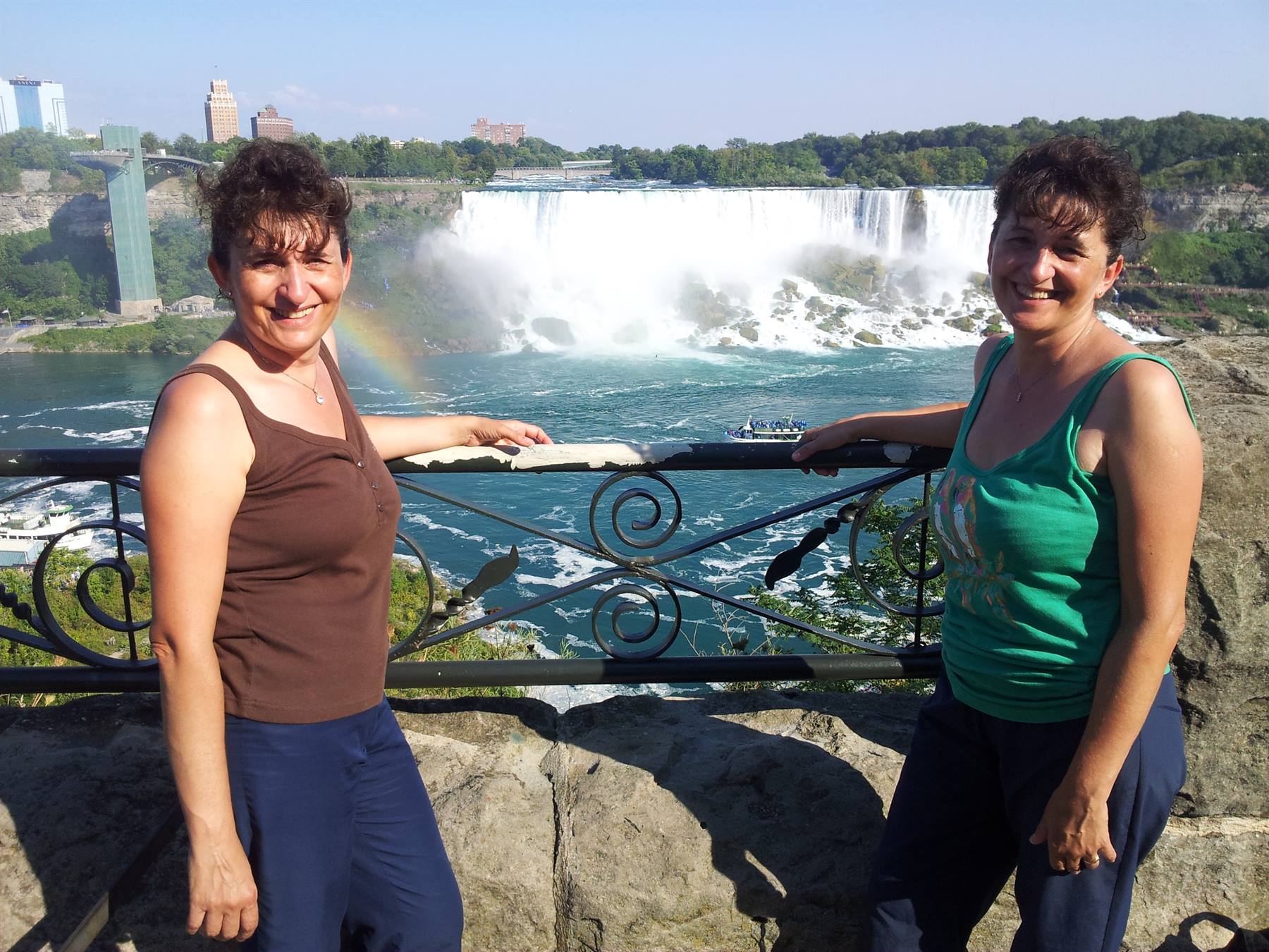 Two women smiling by an iron railing in front of Horseshoe Falls, rainbow and boat visible