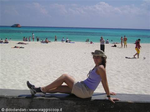 Woman in hat sitting on a wall, white sand beach and crowded turquoise sea