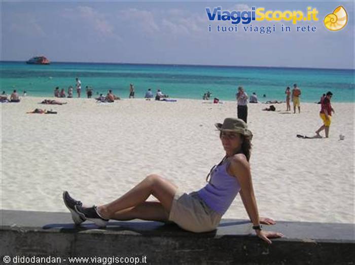 Woman in hat sitting on a wall, white sand beach and crowded turquoise sea
