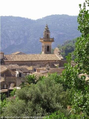 Stone bell tower rising above village rooftops, green trees and mountains, Valldemossa