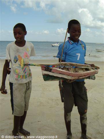 Two boys on a sandy beach, one carrying a small painted wooden boat