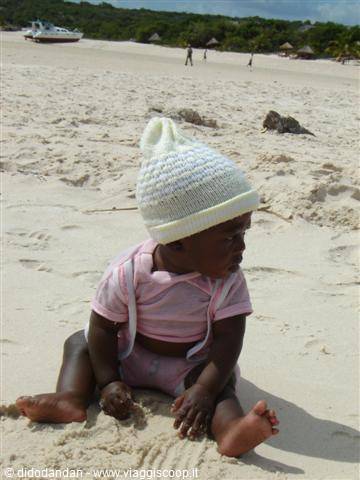 Baby in white knit hat sitting on sand, beach with boats and umbrellas