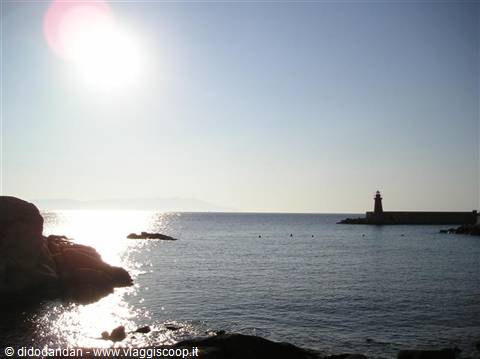 Lighthouse on stone pier, low sun reflecting on sea, rocks in foreground