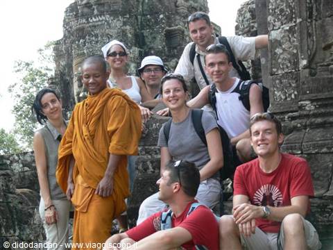 A Buddhist monk in orange robe poses with a group of tourists among stone ruins