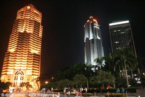 Illuminated skyscrapers at night, palm trees and people strolling in the square