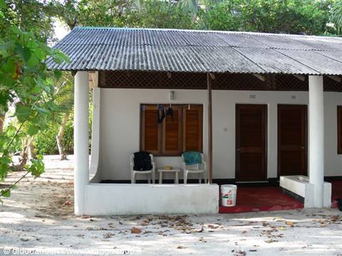 White bungalow with corrugated metal roof, plastic chairs on veranda, sand and tropical trees