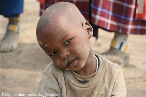 Maasai child with shaved head, wearing worn t-shirt, near adults with colorful beaded anklets