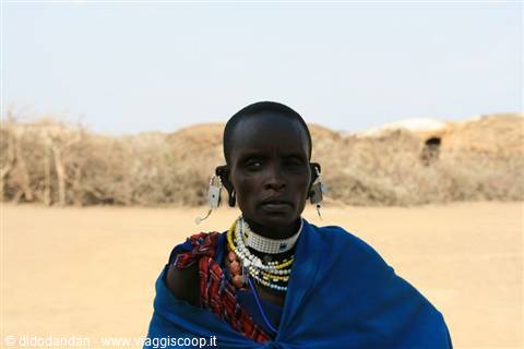 Maasai woman with beaded jewelry and blue shawl in front of huts