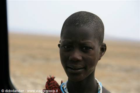 Boy wearing white beaded necklace looking toward vehicle, arid dryland