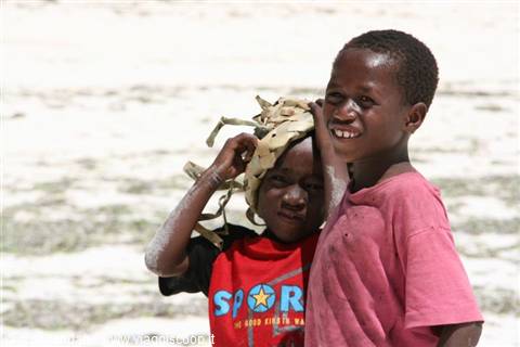 Two smiling children, one wearing a cloth headwrap, on pale sandy beach
