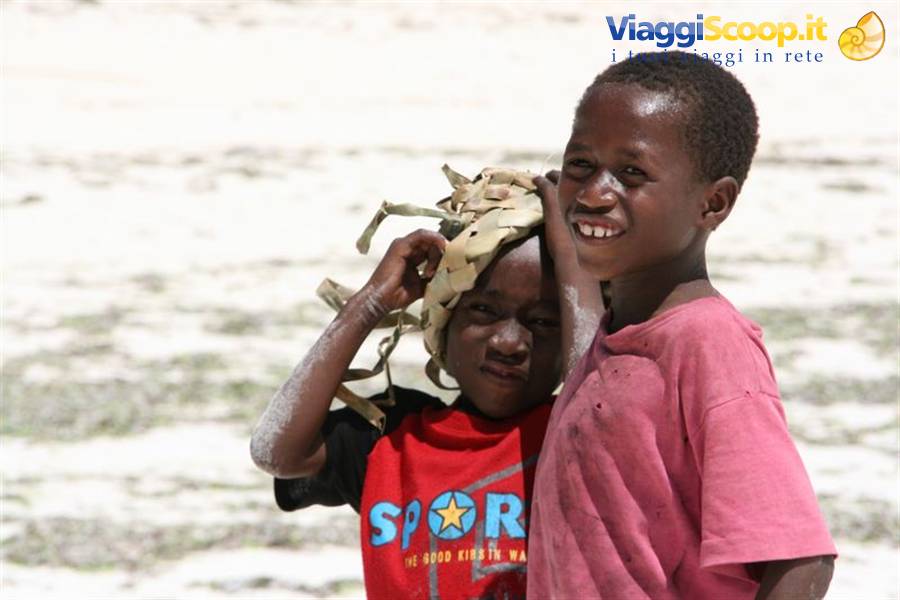 Two smiling children, one wearing a cloth headwrap, on pale sandy beach