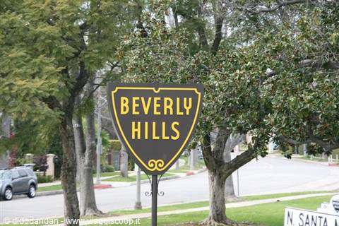 Yellow Beverly Hills shield sign beside a tree-lined street, cars parked under trees