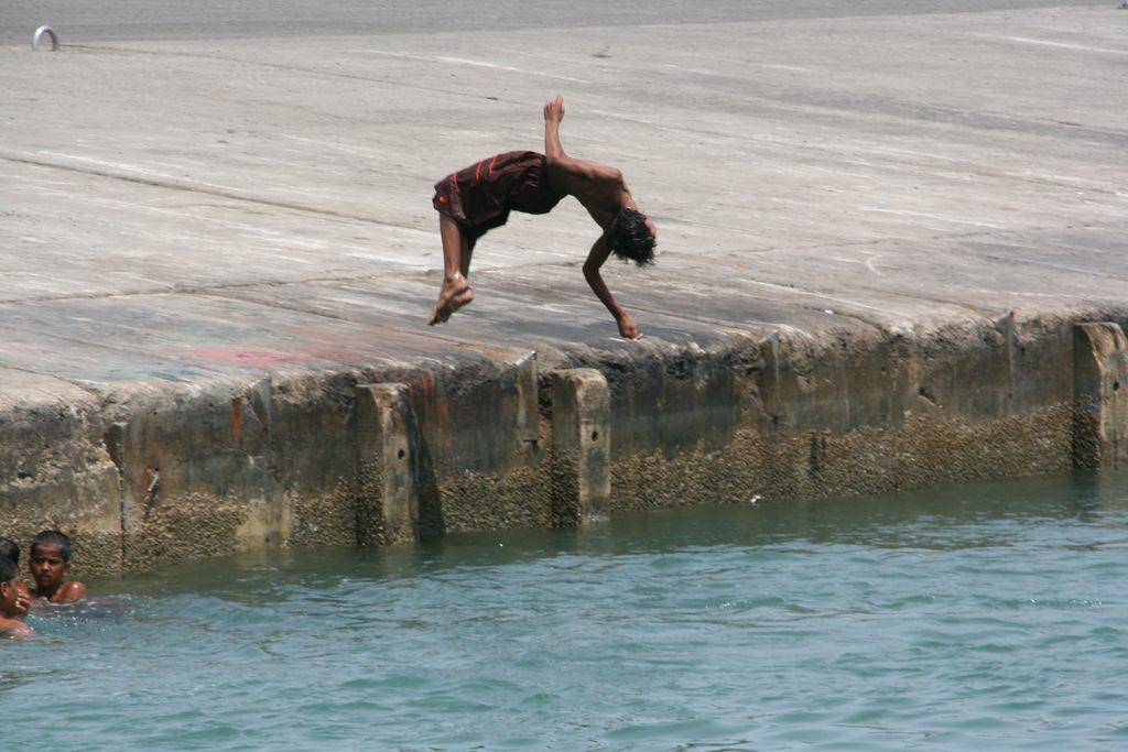 Boy backflipping off the concrete pier, others swimming in the green water